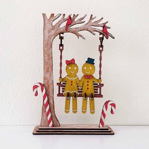 Puede incluir: Decoración navideña de madera con dos hombrecitos de jengibre en un columpio. El columpio está suspendido de un árbol con pájaros rojos. Bastones de caramelo flanquean la base. La pareja de pan de jengibre se da la mano.