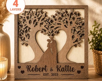 Puede incluir: Un cuadro decorativo cuadrado de madera con cuatro capas cortadas a láser. El diseño muestra a una pareja bajo dos árboles cuyas ramas forman un corazón. El texto "Robert & Kattie" y "EST. 2023" está grabado en la parte inferior.