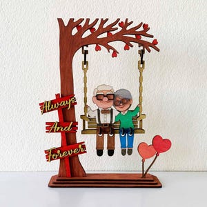 May include: A wooden decorative piece featuring a couple on a swing under a tree with heart-shaped leaves. The words "Always And Forever" are displayed on a sign. Two heart-shaped balloons are also included.