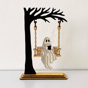 以下が含まれることがあります： 木製のハロウィーンの装飾。コーヒーカップを持ったブランコに乗った、重ねられたゴーストの姿が特徴です。ブランコは木のシルエットから吊り下げられています。ベースは木製で、ゴーストはフレンドリーな表情をしています。