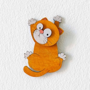 Op de afbeelding: Een platte, oranje houten uitsparing van een cartoonkat met grote, verbaasde ogen, een roze neus en witte pootjes. De kat is afgebeeld in een klimmende of hangende houding met de voor- en achterpoten naar buiten gericht, met een grafisch ontwerp.