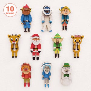 Puede incluir: Una colección de diez adornos de madera con personajes navideños, incluyendo a Santa Claus, un reno, un elfo, un yeti y otras figuras festivas. Cada pieza está pintada con colores vibrantes e incluye un logotipo circular que dice 10 DESIGNS.