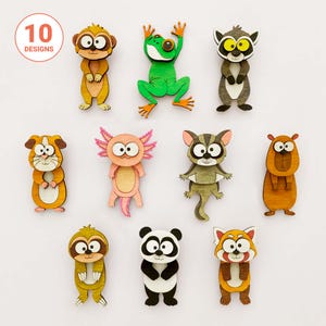May include: A collection of ten wooden animal-shaped ornaments featuring a monkey, frog, lemur, guinea pig, axolotl, sugar glider, capybara, sloth, panda, and red panda, each with large white eyes and a flat, layered design.