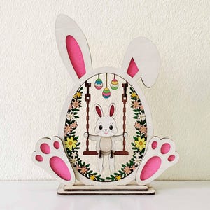 May include: Wooden Easter bunny decoration with a white and pink color scheme. The bunny is sitting on a swing inside an egg-shaped frame, surrounded by flowers. The bunny's ears and paws are pink.