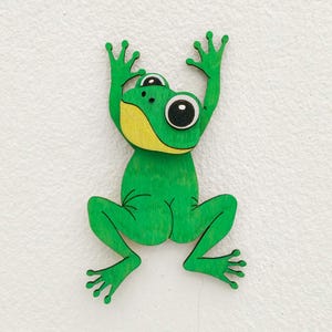 Könnte beinhalten: Ein flacher, grüner Frosch-Ausschnitt aus Holz mit einem gelben Kehlfleck und großen, schwarz-weißen Augen. Der Frosch ist in einer Kletterposition dargestellt, mit erhobenen Vorderbeinen und gespreizten Hinterbeinen, inklusive Holzmaserung.