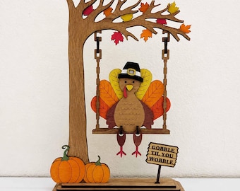 Può includere: Una decorazione in legno con un tacchino che indossa un cappello da pellegrino nero seduto su un'altalena appesa a un ramo. L'albero ha foglie autunnali colorate, due zucche arancioni e un cartello con la scritta GOBBLE TIL YOU WOBBLE.