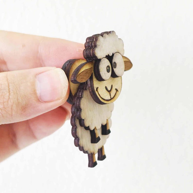 May include: A wooden sheep-shaped brooch with a smiling face and large eyes. The brooch is made of layered wood, with a light brown body and a darker brown outline. The sheep's wool is a lighter color, and the brooch is held in a hand.
