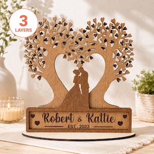 Peut inclure: Un ornement en bois découpé au laser à trois couches représentant deux arbres formant un cœur avec un couple debout au centre. La base rectangulaire est gravée avec le texte Robert & Kattie et EST. 2023, ornée de quatre petits cœurs.