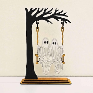 May include: A wooden Halloween decoration featuring two white ghost figures sitting on a swing. The swing is suspended from a black tree silhouette. The base is made of layered wood.