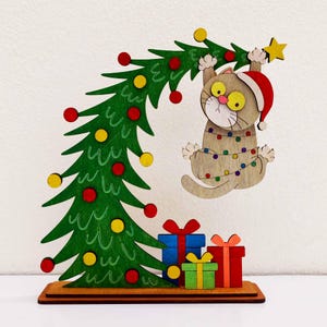 May include: A wooden Christmas decoration featuring a green tree with red and yellow ornaments, a tan cat wearing a red Santa hat tangled in colorful string lights, and three stacked gift boxes in blue, green, and red at the base.