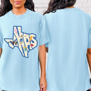 May include: A light blue t-shirt featuring a graphic of the state of Texas. The graphic is made up of the letters "TEXAS" and includes various icons representing Texan culture, such as a cowboy, cotton candy, a hot dog, and a carousel.