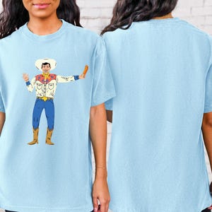 May include: A light blue t-shirt featuring a cartoon illustration of a cowboy holding a drumstick. The cowboy is wearing a white shirt with a red and blue waistcoat, blue jeans, and a cowboy hat.