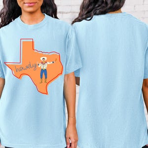 以下が含まれることがあります： 薄い青色のTシャツに、白いシャツと青いジーンズを着たカウボーイの漫画のイラストが描かれている。カウボーイはテキサス州の輪郭の中に立っている。テキサス州の輪郭の中に「howdy」という言葉が筆記体で書かれている
