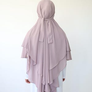 Könnte beinhalten: Ein mauvefarbenes Hijab mit einem mehrschichtigen Design. Das Hijab ist mit einer Schleife am Hinterkopf gebunden.