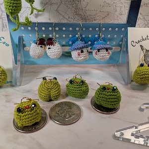 May include: Crocheted earrings in the shape of green frogs and white mushrooms with blue hats. The earrings are displayed on a white grid background with a sign that reads "Crochet with Meow".