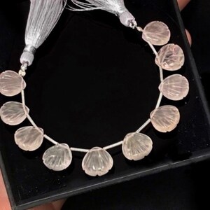Peut inclure: Un collier de perles de quartz rose, chacune sculptée en forme de coquille. Les perles sont enfilées sur un fil d'argent et ont un pompon à chaque extrémité.