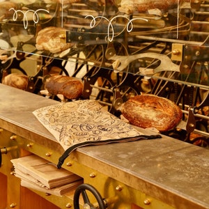 May include: A close-up of a bakery display featuring a selection of freshly baked loaves of bread. The bread is arranged on a metal rack with a decorative pattern. The rack is made of metal and has a gold finish. The bread is a light brown colour and has a crusty texture.