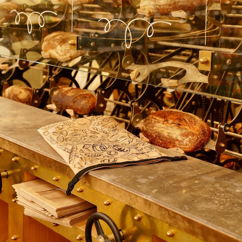 May include: A close-up of a bakery display featuring a selection of freshly baked loaves of bread. The bread is arranged on a metal rack with a decorative pattern. The rack is made of metal and has a gold finish. The bread is a light brown colour and has a crusty texture.