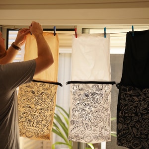 May include: Four cloth bags hanging on a washing line. The bags are made of different coloured fabrics: beige, white, and black. Each bag has a black and white illustration of various types of bread on the front.