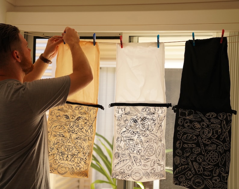 May include: Four cloth bags hanging on a washing line. The bags are made of different coloured fabrics: beige, white, and black. Each bag has a black and white illustration of various types of bread on the front.