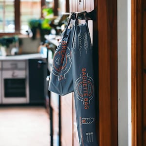 May include: A gray reusable baguette bag with the text "Baguette Bag" and "Reusable" printed on it. The bag is hanging on a door handle.