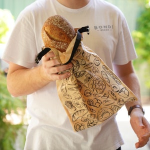 May include: A person holding a brown reusable bread bag with a black strap and a white background with a black print of various bread designs. The bag says "BONDI BEACH" in black text.
