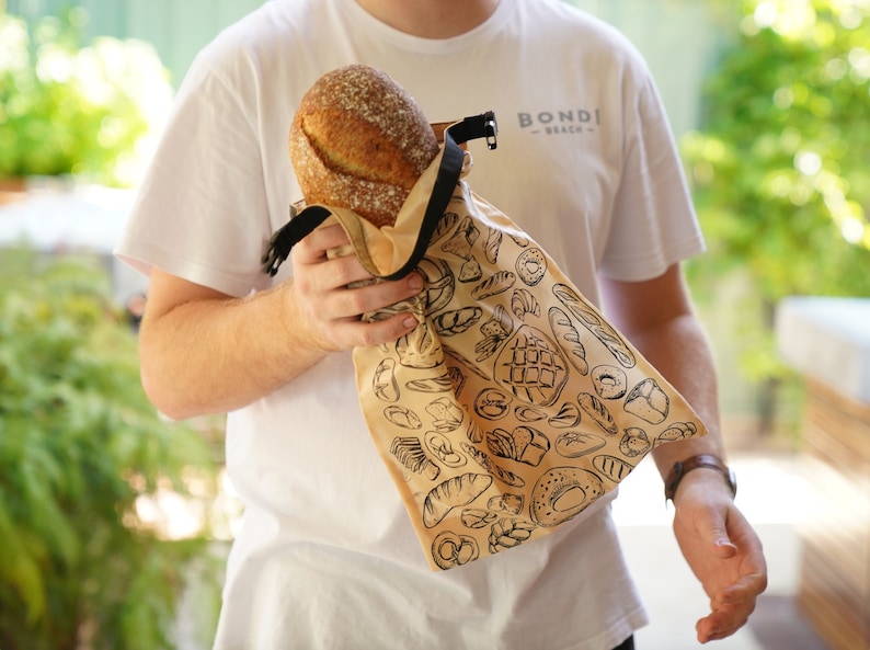 May include: A person holding a brown reusable bread bag with a black strap and a white background with a black print of various bread designs. The bag says "BONDI BEACH" in black text.