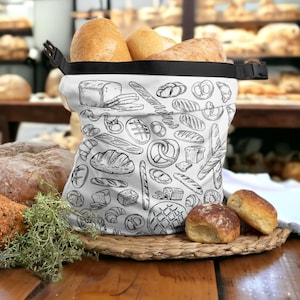 May include: A white canvas bag with a black zipper and a black handle. The bag is filled with various types of bread, including loaves, rolls, and baguettes. The bag is printed with a black and white pattern of various bread items, including pretzels, croissants, and loaves.