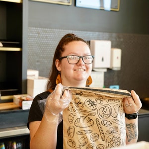 May include: A person wearing a black shirt and a brown paper bag with a black and white illustration of various breads and pastries. The bag has a black strap.