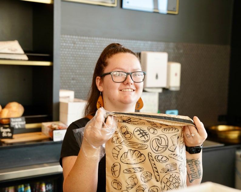 May include: A person wearing a black shirt and a brown paper bag with a black and white illustration of various breads and pastries. The bag has a black strap.