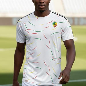 Könnte beinhalten: Weißes Fußballtrikot mit roten, grünen und gelben geschwungenen Linien. Das Trikot hat ein Kamerun-Flaggenemblem und schwarze Akzente auf den Schultern. Die Person trägt weiße Shorts.