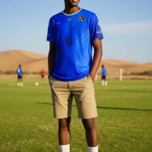 Peut inclure: Une personne porte un maillot de foot bleu avec le numéro 6, un short kaki, des chaussettes blanches et des chaussures noires sur un terrain vert. D'autres personnes jouent au football en arrière-plan.
