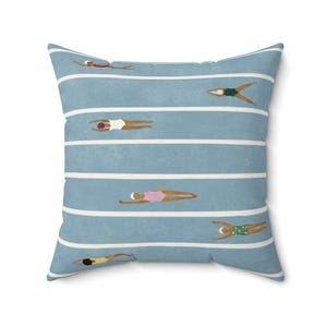 Pode incluir: Uma almofada quadrada com fundo azul claro e riscas horizontais brancas. O design inclui várias figuras ilustradas a nadar com fatos de banho de cores diferentes, incluindo vermelho, verde, branco, rosa e amarelo, todas com toucas de natação brancas.