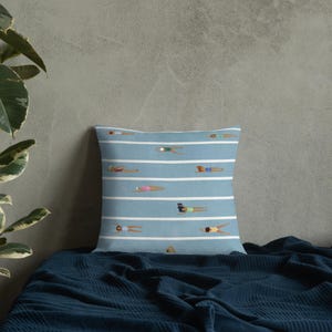 Peut inclure: Un coussin carré avec un fond bleu clair et des rayures horizontales blanches. Le motif présente plusieurs petites illustrations de personnes nageant dans divers maillots de bain colorés, placées entre les rayures blanches.