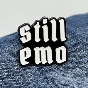 May include: A black and white enamel pin with the words "still emo" in a bold, stylized font. The pin is rectangular with rounded corners and is set against a blue denim background. The text is in white, with a black border.