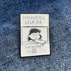 May include: A rectangular enamel pin with a black border. The pin features a black and white cartoon image of a character in a booth with the text "PSYCHIATRIC HELP 5¢" and "THE DOCTOR IS IN". The pin is set against a blue denim background.