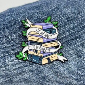 May include: A stack of books with a white ribbon wrapped around them. The ribbon has the text "Ask me about my to be read pile" on it. The books are purple, blue, and yellow. The pin is on a blue denim background.