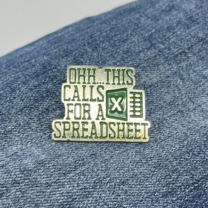 May include: A rectangular enamel pin with the text "OHH THIS CALLS FOR A SPREADSHEET" in green and white. The pin features a green spreadsheet icon. The pin is set against a blue denim background.