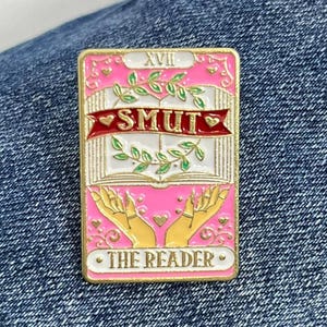 May include: A pink and gold enamel pin featuring a tarot card design. The card is labelled "XVII" and "The Reader". The card depicts a book with a heart and the word "SMUT" on a red banner.