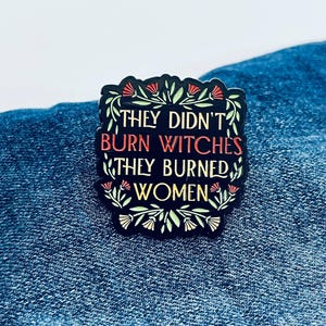 May include: Black enamel pin with the text "THEY DIDN'T BURN WITCHES THEY BURNED WOMEN" in red and white. The pin is framed by green leaves and red and white flowers. The pin is on a blue denim background.