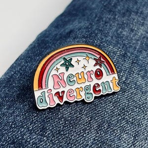 May include: A rainbow enamel pin with the text "Neuro divergent" in white letters with pink, red, and green outlines. The rainbow has a yellow, orange, red, pink, and blue colour scheme. There are five gold stars on the pin.