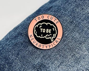以下が含まれることがあります： 「TOO CUTE TO BE NEUROTYPICAL」の文字が入った丸いエナメルピン。ピンは、白い脳のグラフィックとテキストが入った黒い中心部、ピンクのリングと白いテキストで囲まれ、青いデニムの背景に配置されています。