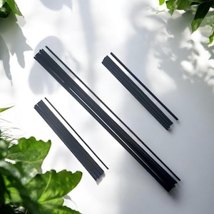 May include: A collection of black diffuser reeds, presented on a white background with leafy shadows. The reeds, designed for fragrance diffusion, are thin and cylindrical. The image is a top-down view, showcasing the product.
