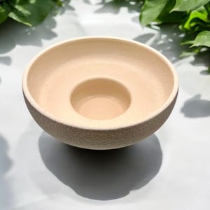 May include: A beige ceramic candle holder with a textured exterior. The holder has a circular shape with a central well for a candle. The design is simple and modern, suitable for home decor.