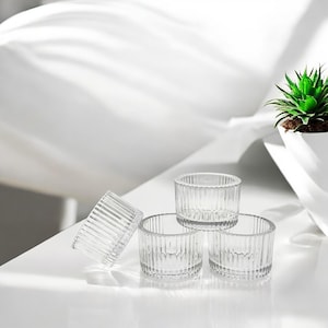 May include: Four clear glass votive candle holders with vertical ribbed detailing. One holder is tilted, and the others are stacked on a white surface. A small potted succulent plant is in the background.