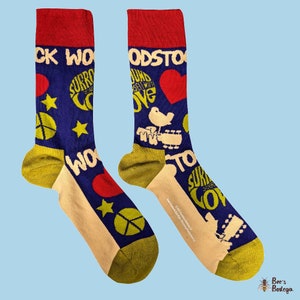 以下が含まれることがあります： 赤、白、黄色のデザインの青と黄色の靴下1足。靴下には「ウッドストック」と「愛で包まれ」の文字と、平和の象徴、星、鳥が描かれています。