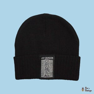 Puede incluir: Gorro negro con un parche rectangular con el texto "JOY DIVISION" encima de un gráfico en blanco y negro y las palabras "UNKNOWN PLEASURES" debajo. El gorro está hecho de un material de punto suave.