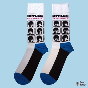 Op de afbeelding: Paar witte sokken met zwarte en blauwe accenten. Elke sok heeft een raster van zwart-wit afbeeldingen van The Beatles en de tekst "A Hard Day's Night". De sokken hebben een blauwe teen en hiel.