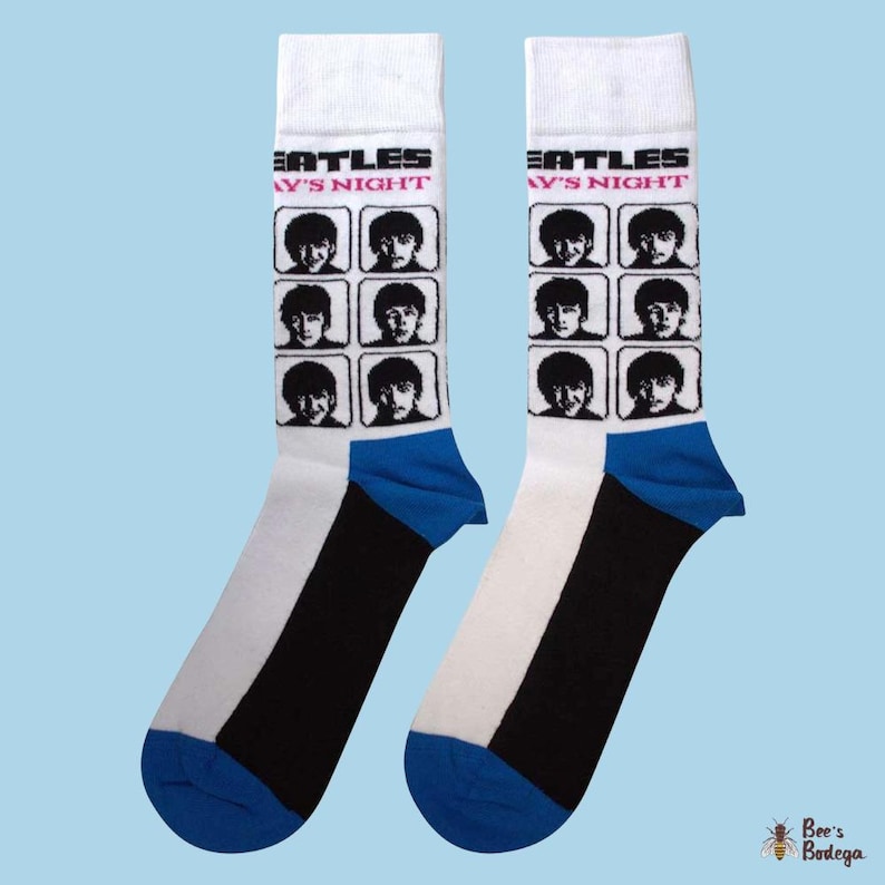 Op de afbeelding: Paar witte sokken met zwarte en blauwe accenten. Elke sok heeft een raster van zwart-wit afbeeldingen van The Beatles en de tekst "A Hard Day's Night". De sokken hebben een blauwe teen en hiel.
