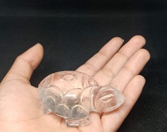 May include: A clear quartz crystal carved into the shape of a turtle. The turtle is sitting on a hand.